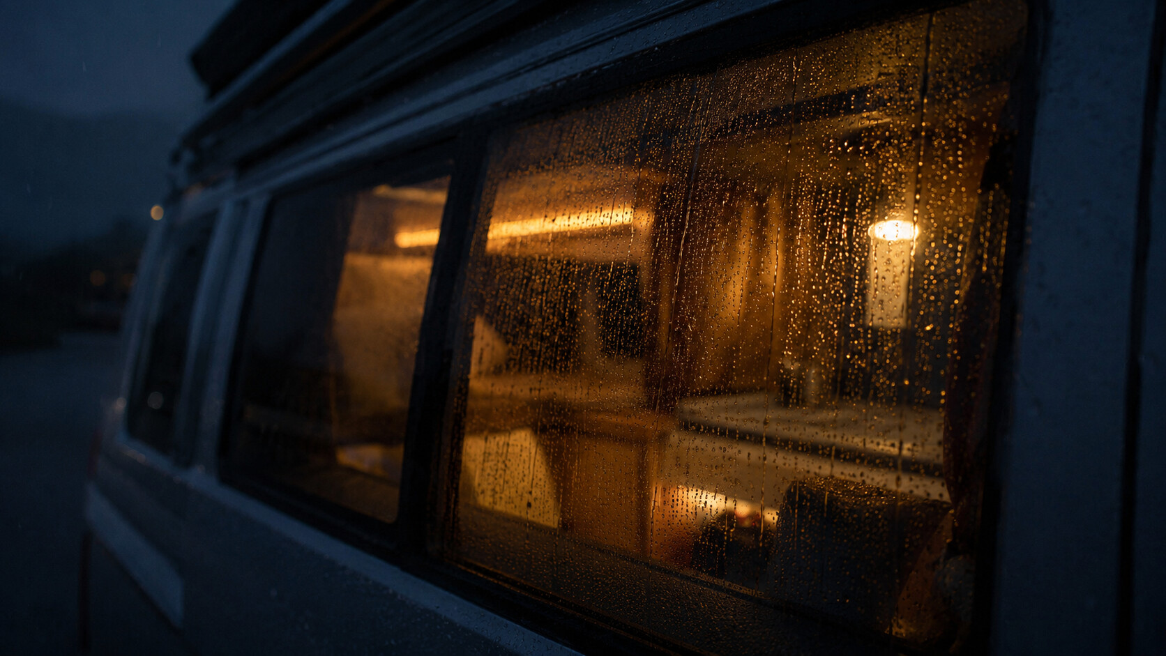 Regentropfen auf dem Fenster eines Campingbusses bei Nacht mit warmem Licht im Inneren