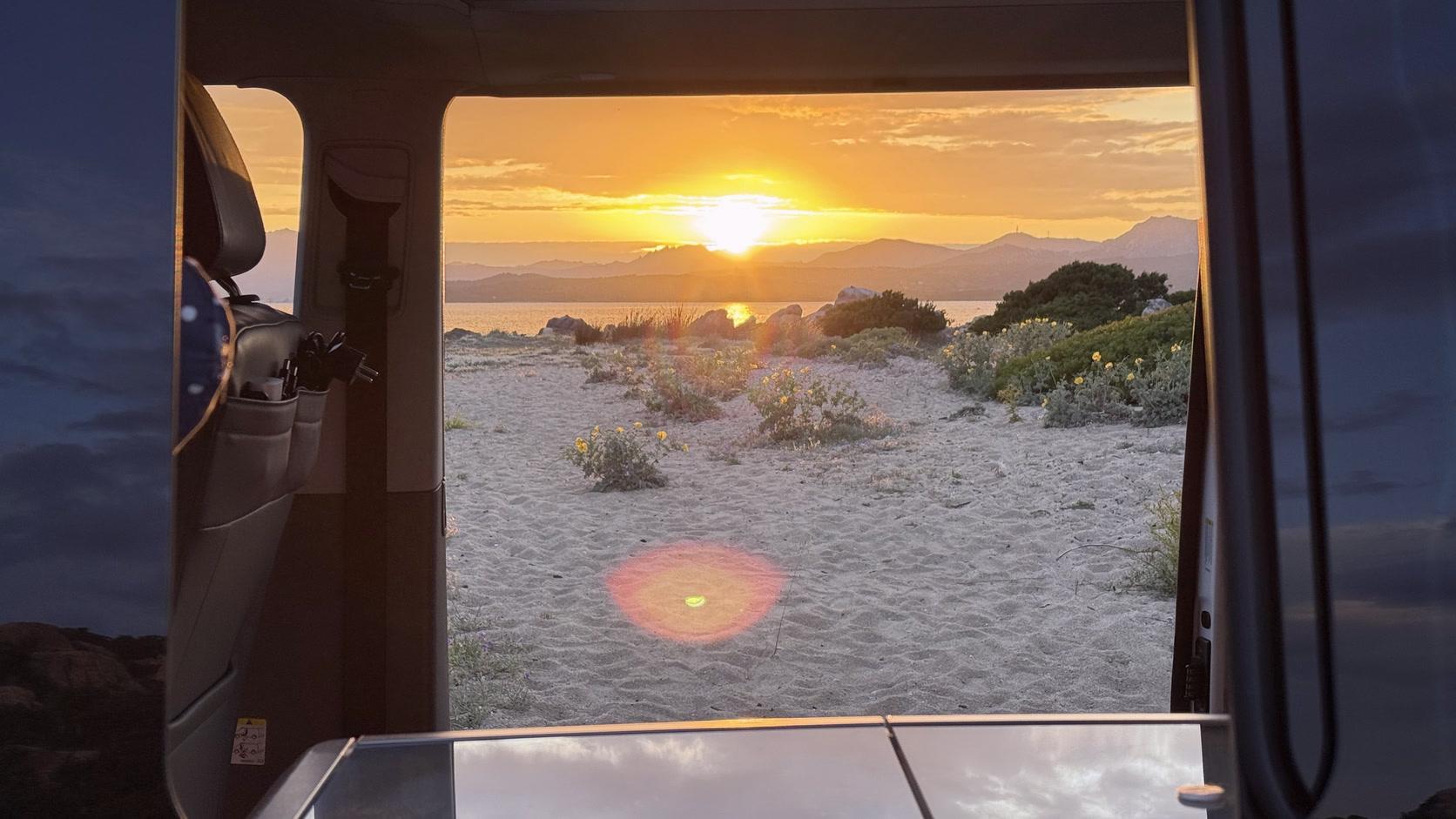 Blick aus der offenen Schiebetür über Dünen auf das Meer, die Sonne steht tief.